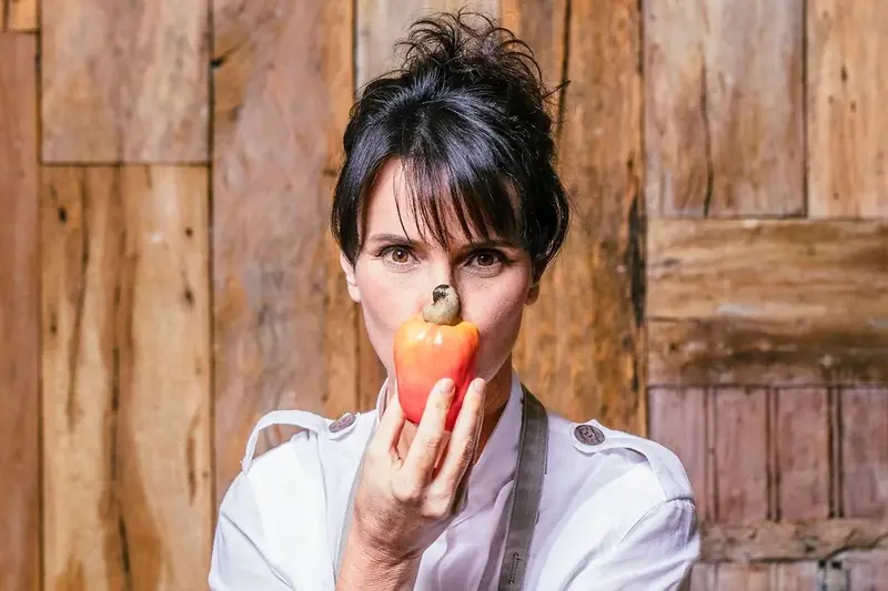 Retrato da chef Renata Vanzetto segurando um caju diante do rosto, com parede de madeira ao fundo